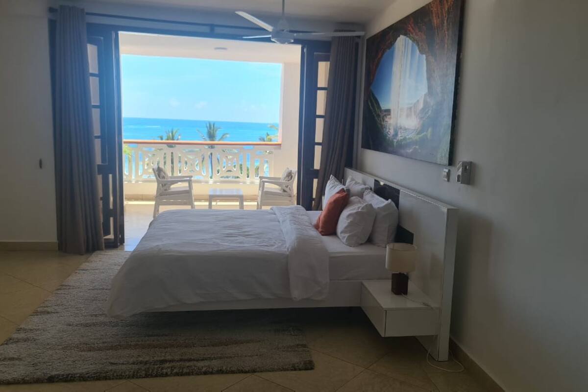 A bedroom with a bed, a painting, and a view of the ocean through an open door.