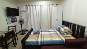 A bedroom with a bed, a desk, a chair, a TV, a fan, and a wardrobe.