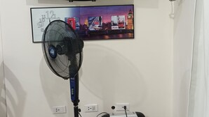 A room with a flat-screen TV mounted on the wall, a standing fan, and a heater on the floor.