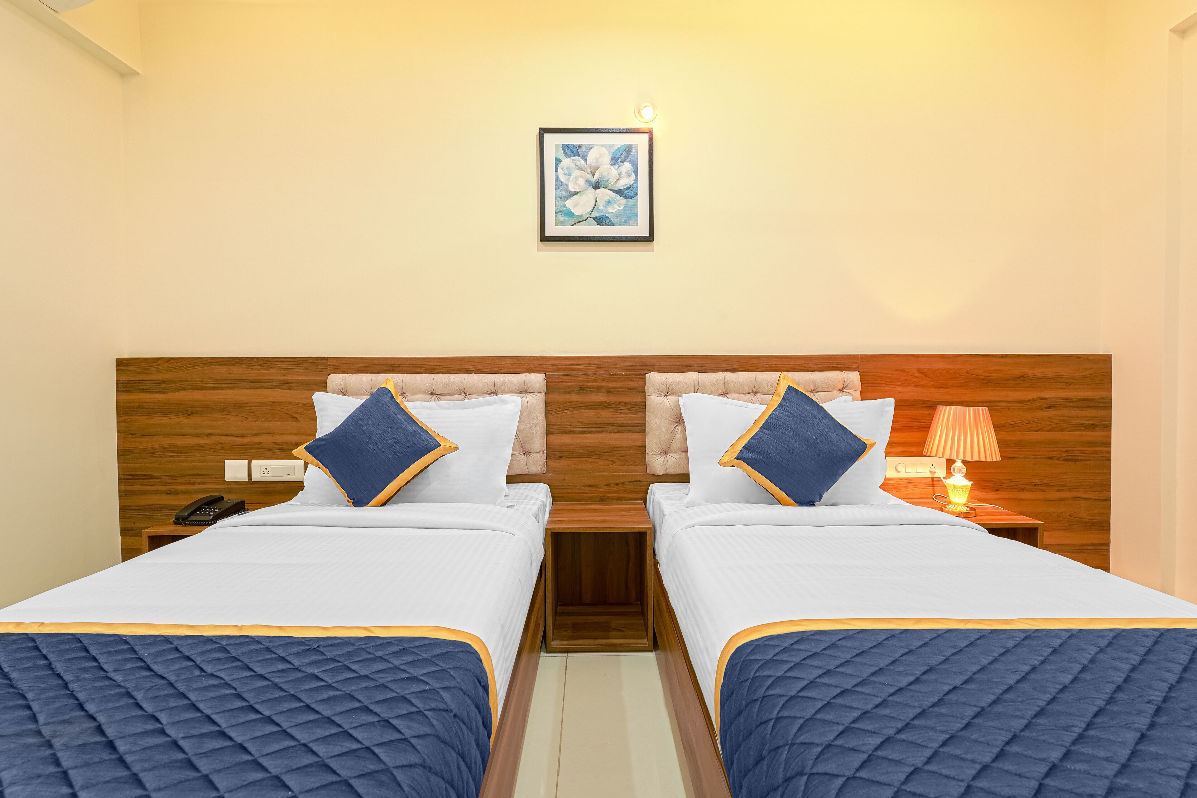 A hotel room with two beds, a wooden headboard, a nightstand with a lamp, and a framed artwork on the wall.
