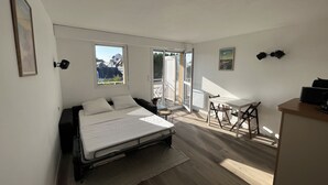 Une chambre avec un lit, un bureau et une chaise. Il y a une fenêtre donnant sur des immeubles et une porte qui mène à un espace extérieur.