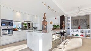Cozinha moderna com armários brancos, uma ilha central e uma área de jantar com mesa e cadeiras brancas.
