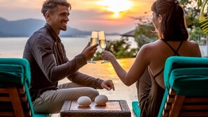 Duas pessoas brindando com taças de champanhe em uma mesa com dois ovos decorativos, com vista para um corpo d'água e montanhas.
