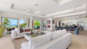 Ampla sala de estar com um sofá branco grande, uma mesa de centro com livros e objetos decorativos, e vista para o oceano através de grandes janelas.