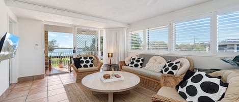 Ein Wohnzimmer mit einem runden Couchtisch, Korbssofas mit gepunkteten Kissen und Blick auf ein Gewässer durch eine Glastür.