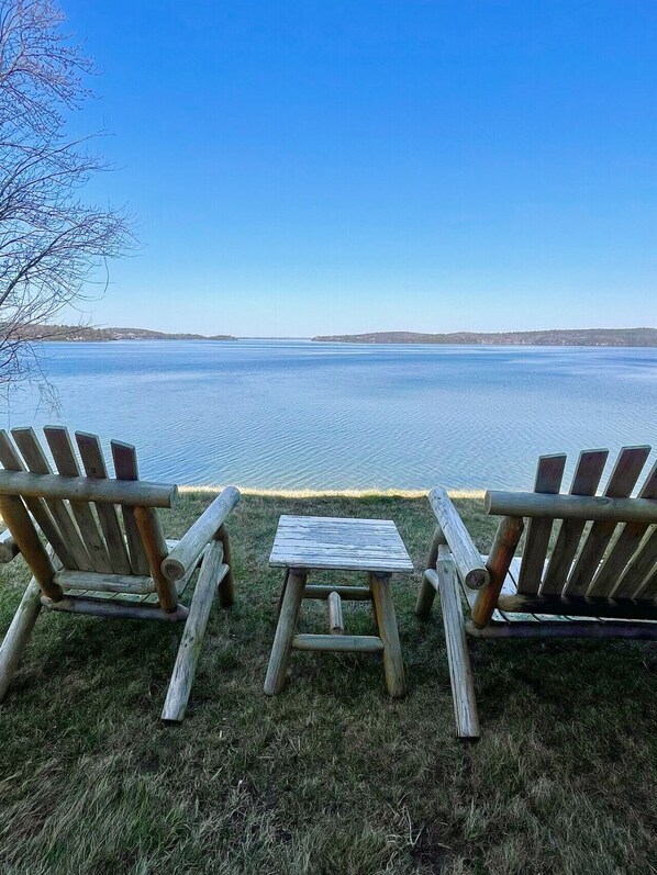 Two wooden Adirondack chairs facing a calm lake with a clear sky.