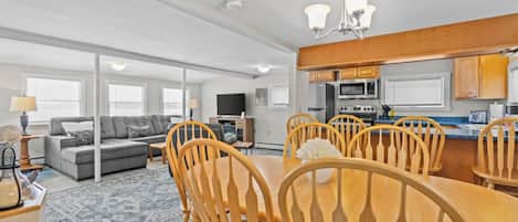 A modern living room with a sofa, coffee table, and dining table with chairs. There is a kitchen area with cabinets and appliances.