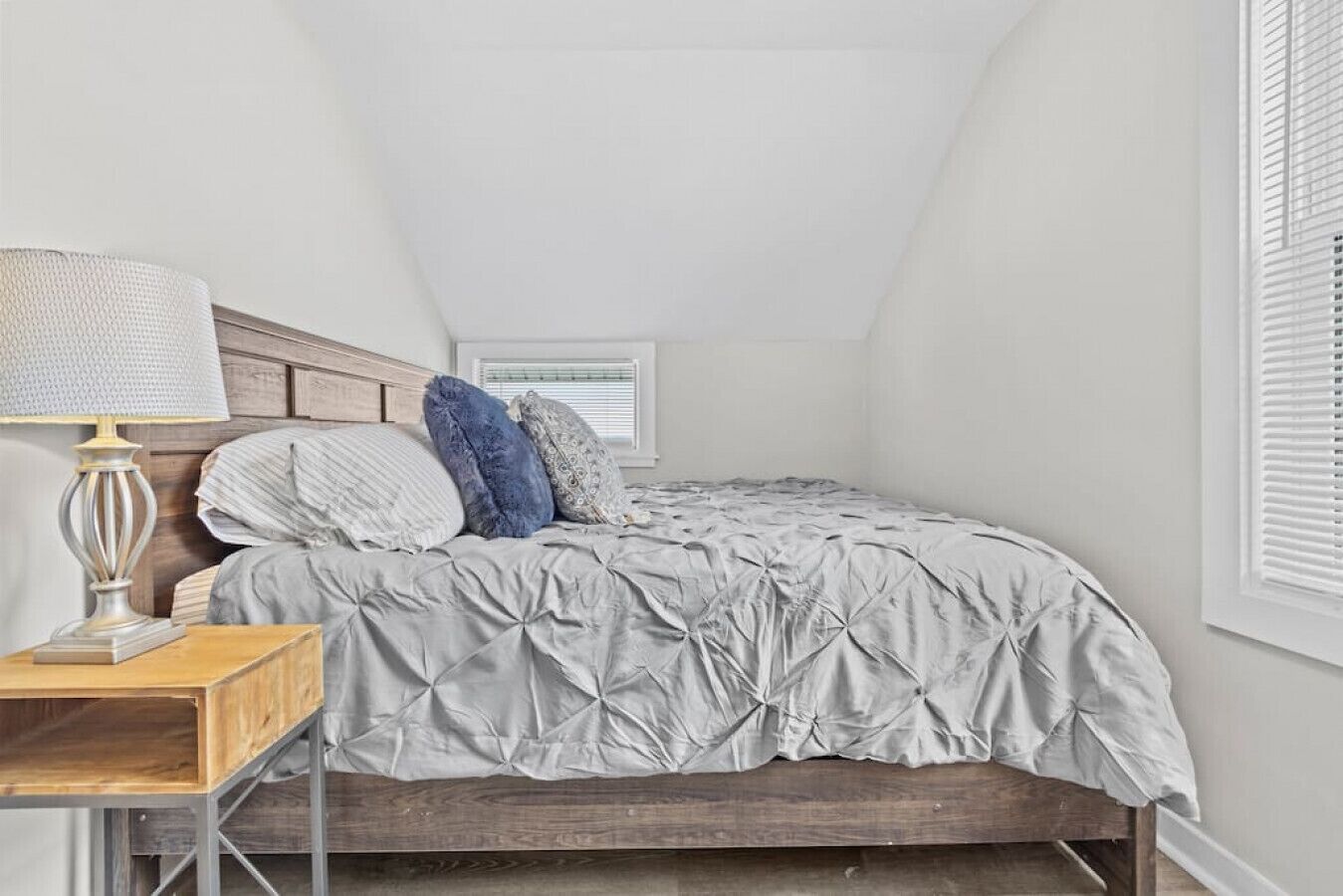 A bedroom with a sloped ceiling, a bed with a grey duvet, a wooden nightstand with a lamp, and a window with blinds.