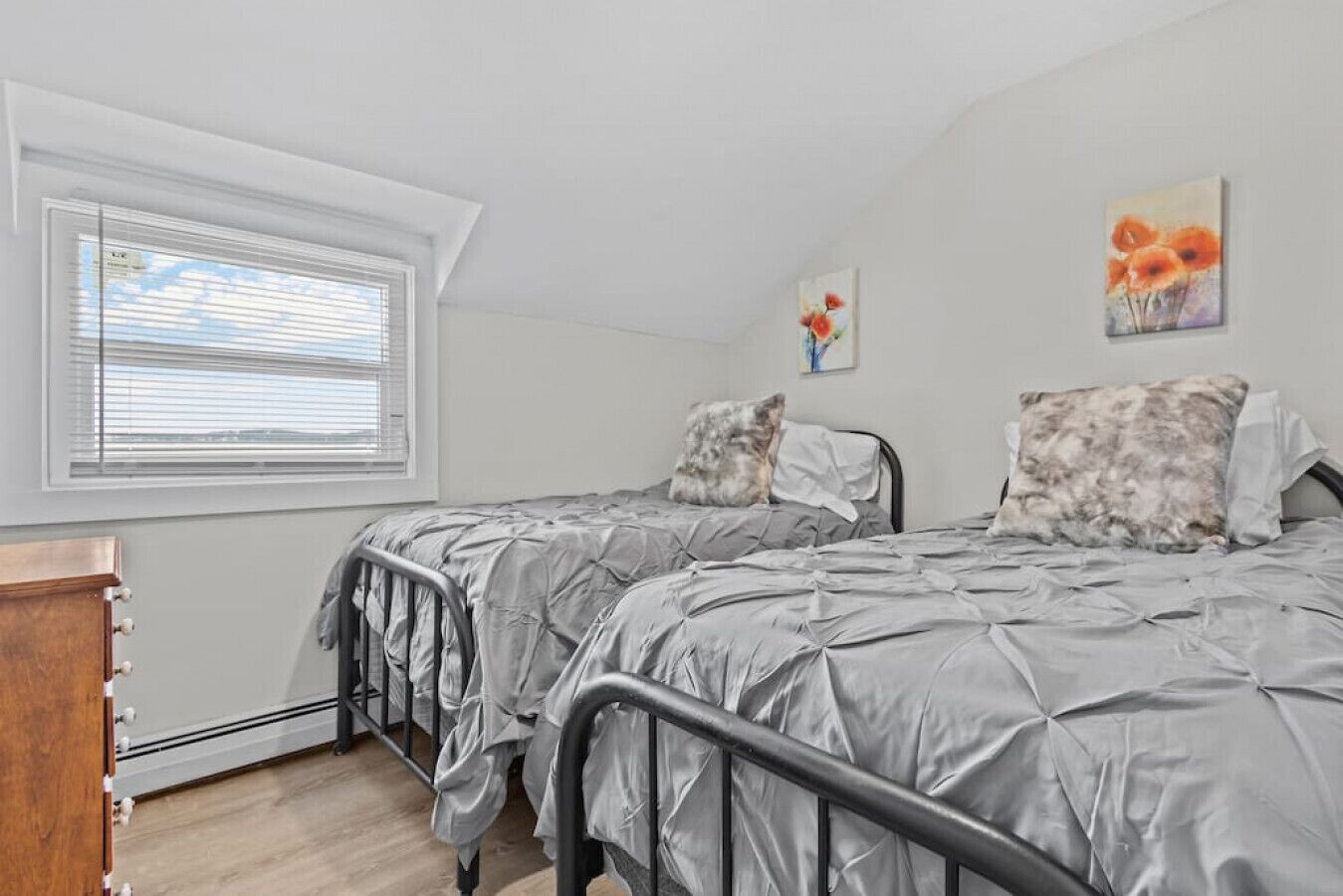 A bedroom with two beds, a window with blinds, a wooden dresser, and two paintings on the wall.