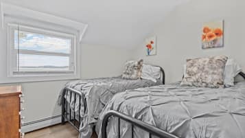 A bedroom with two beds, a window with blinds, a wooden dresser, and two paintings on the wall.