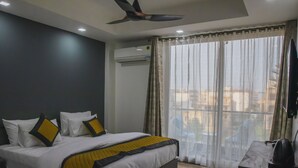 A hotel room with a bed, a ceiling fan, an air conditioning unit, a window with curtains, and a desk with a kettle. - Dev Royale Hotel (Gurugram)