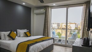 A hotel room with a bed, a ceiling fan, a large window with curtains, and a view of buildings outside. - Dev Royale Hotel (Gurugram)