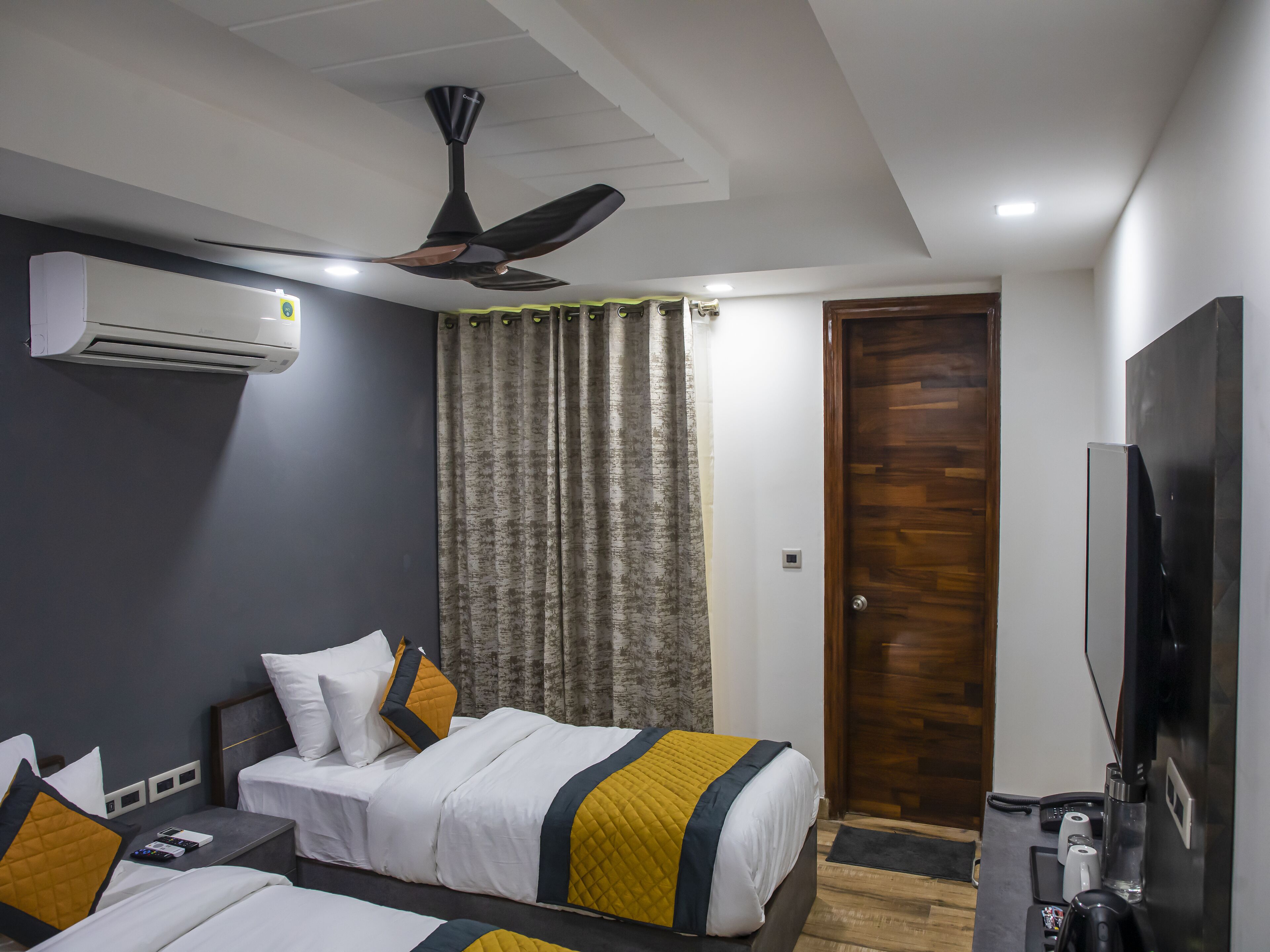 Ein Hotelzimmer mit zwei Betten, einem Deckenventilator, einer Klimaanlage, einer Holztür und einem Flachbildfernseher.