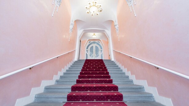 A grand staircase with a red carpet, white railings, and a chandelier at the top. - Beautiful Mala Strana Studio by EasyBNB Premium (Prague)