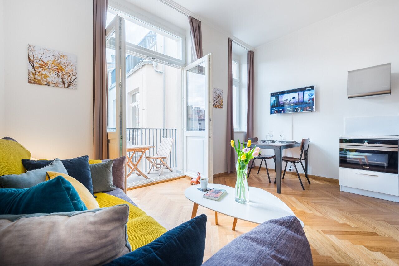 A modern living room with a sofa, coffee table, and a dining area with chairs. There is a TV mounted on the wall and a window with curtains.