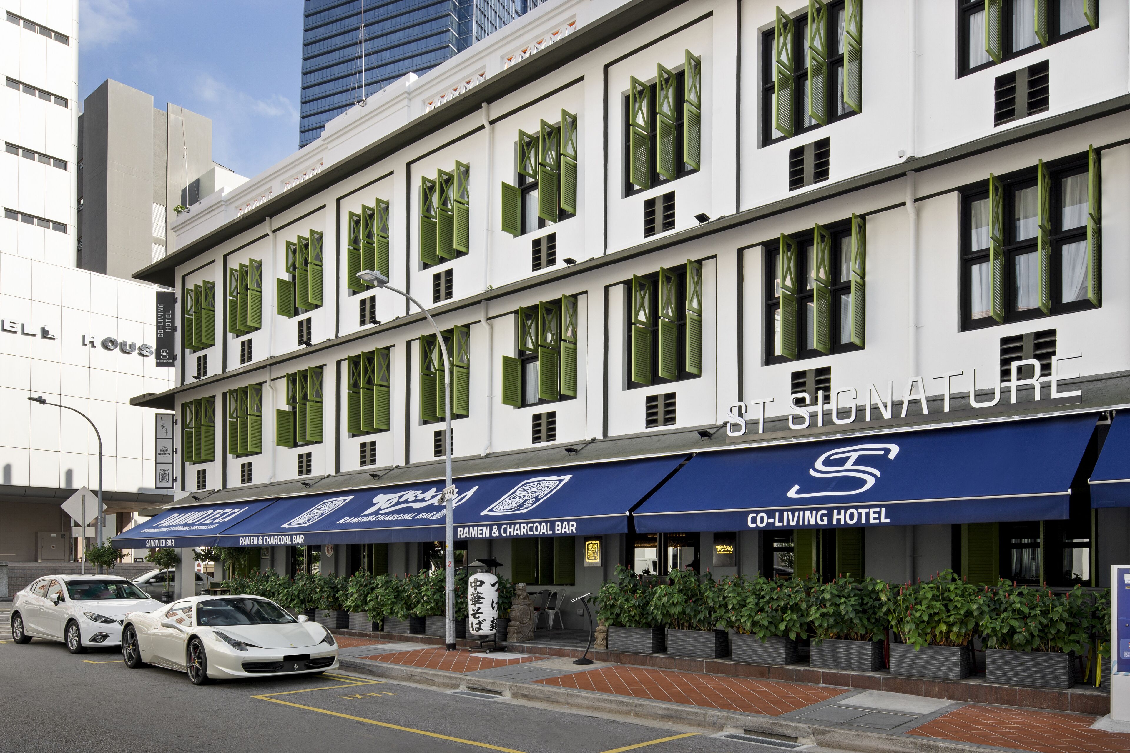 Un hotel con un letrero que anuncia un bar de ramen y carbón, un área de estacionamiento y dos coches blancos estacionados al frente.