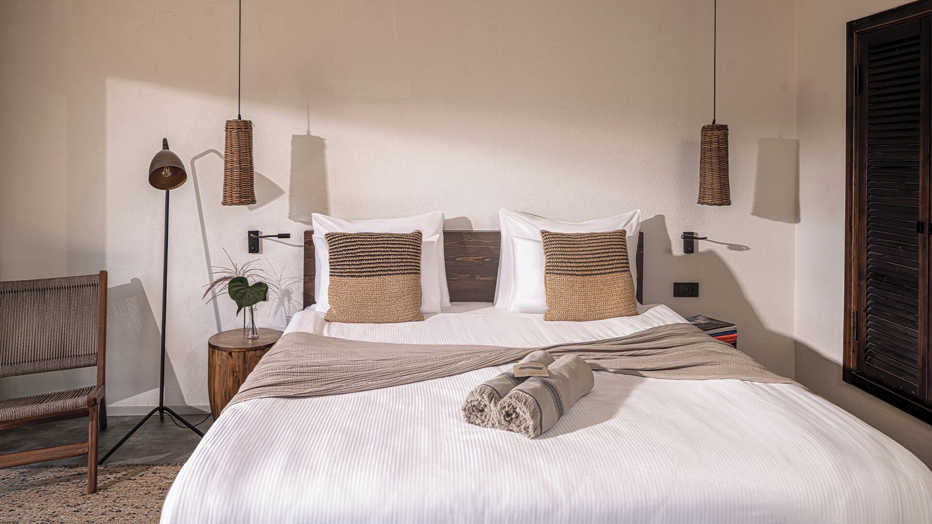 Uma cama bem arrumada com uma toalha enrolada, uma mesinha de vime com uma planta e uma cadeira.