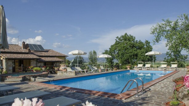 Ein Poolbereich mit Liegestühlen, Sonnenschirmen und einem Gebäude mit Steinbau und Ziegeldach.
