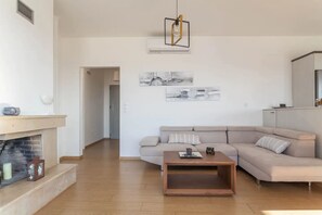 A modern living room with a beige sofa, a wooden coffee table, and a fireplace. There are two wall-mounted artworks and a hanging light fixture.