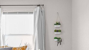 Un dormitori amb llit, una tauleta de nit, una planta penjant i una finestra amb cortines.