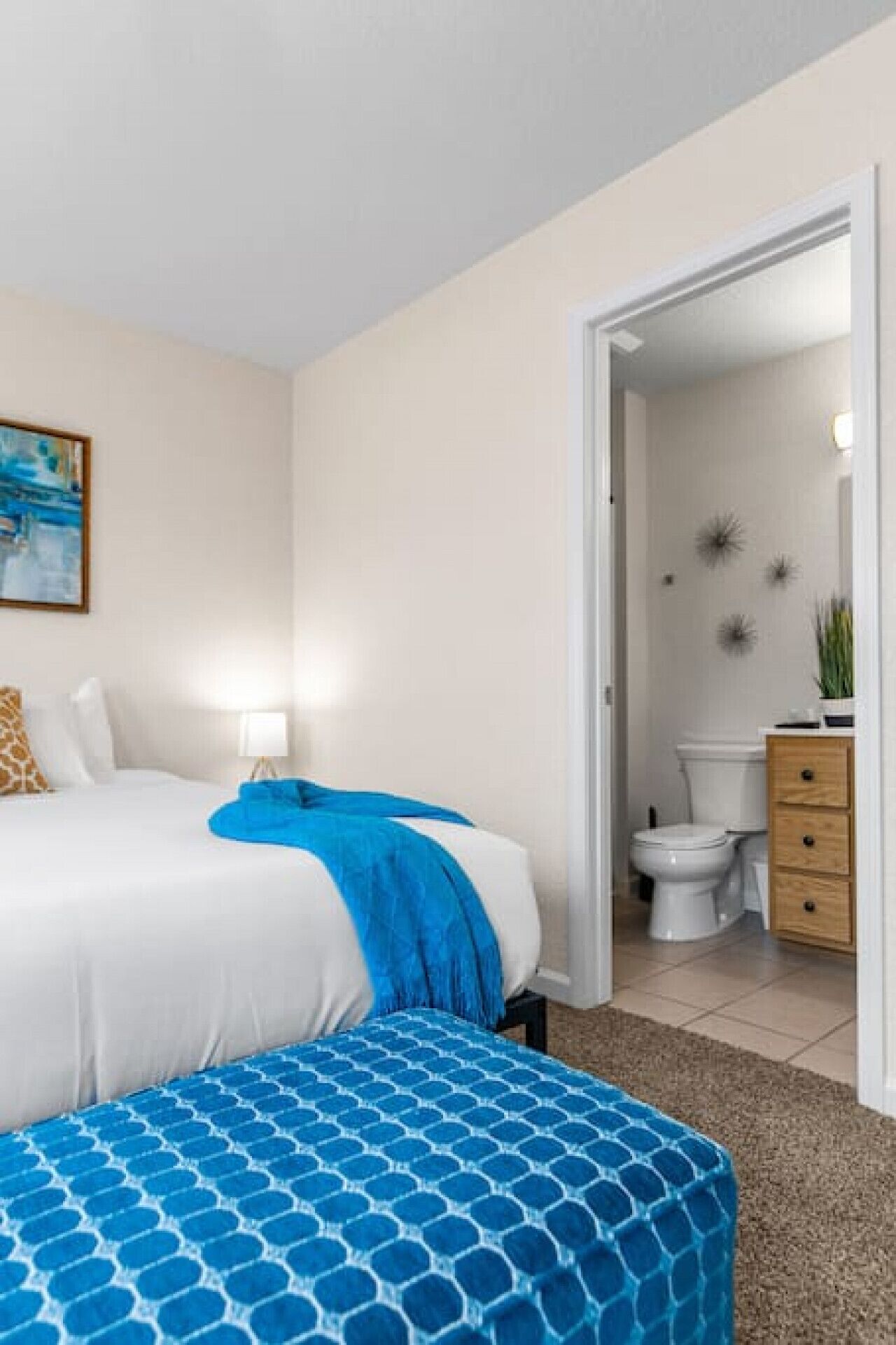 A hotel room with a bed, a bench, a bathroom with a toilet and a sink, and a door leading to another room.