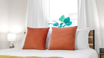 A neatly made bed with white and orange pillows, a yellow throw blanket, and a window with white curtains.
