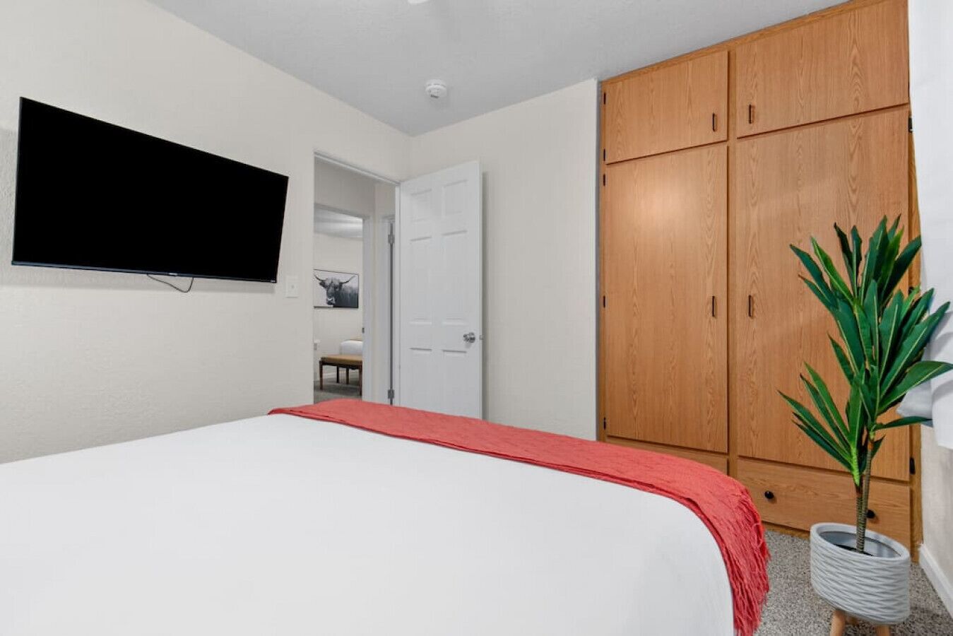 A bedroom with a bed, a TV mounted on the wall, a wooden wardrobe, and a potted plant.
