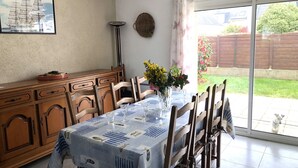 Een eetgedeelte met een tafel gedekt voor vier personen, houten stoelen, een vaas met bloemen en een schilderij van een schip aan de muur.