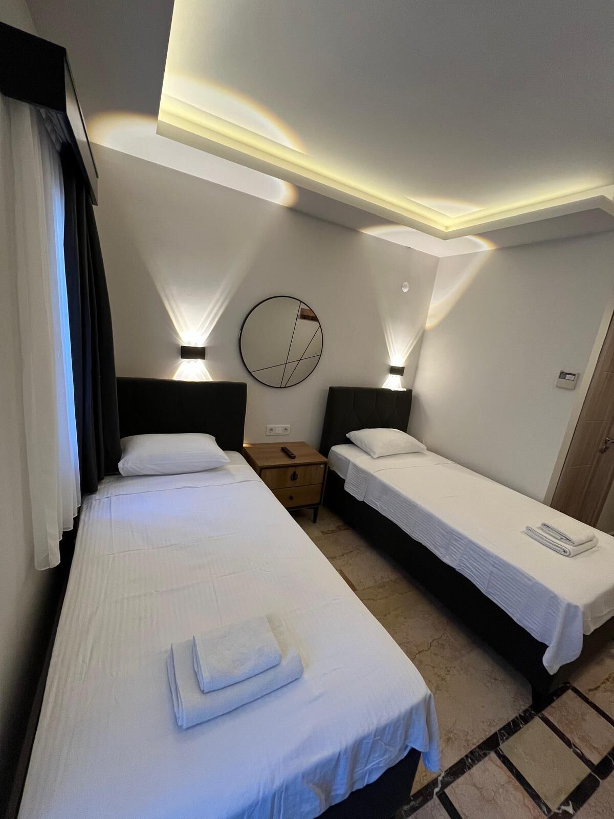 Una habitación de hotel con dos camas individuales, un espejo redondo en la pared y dos lámparas de noche.