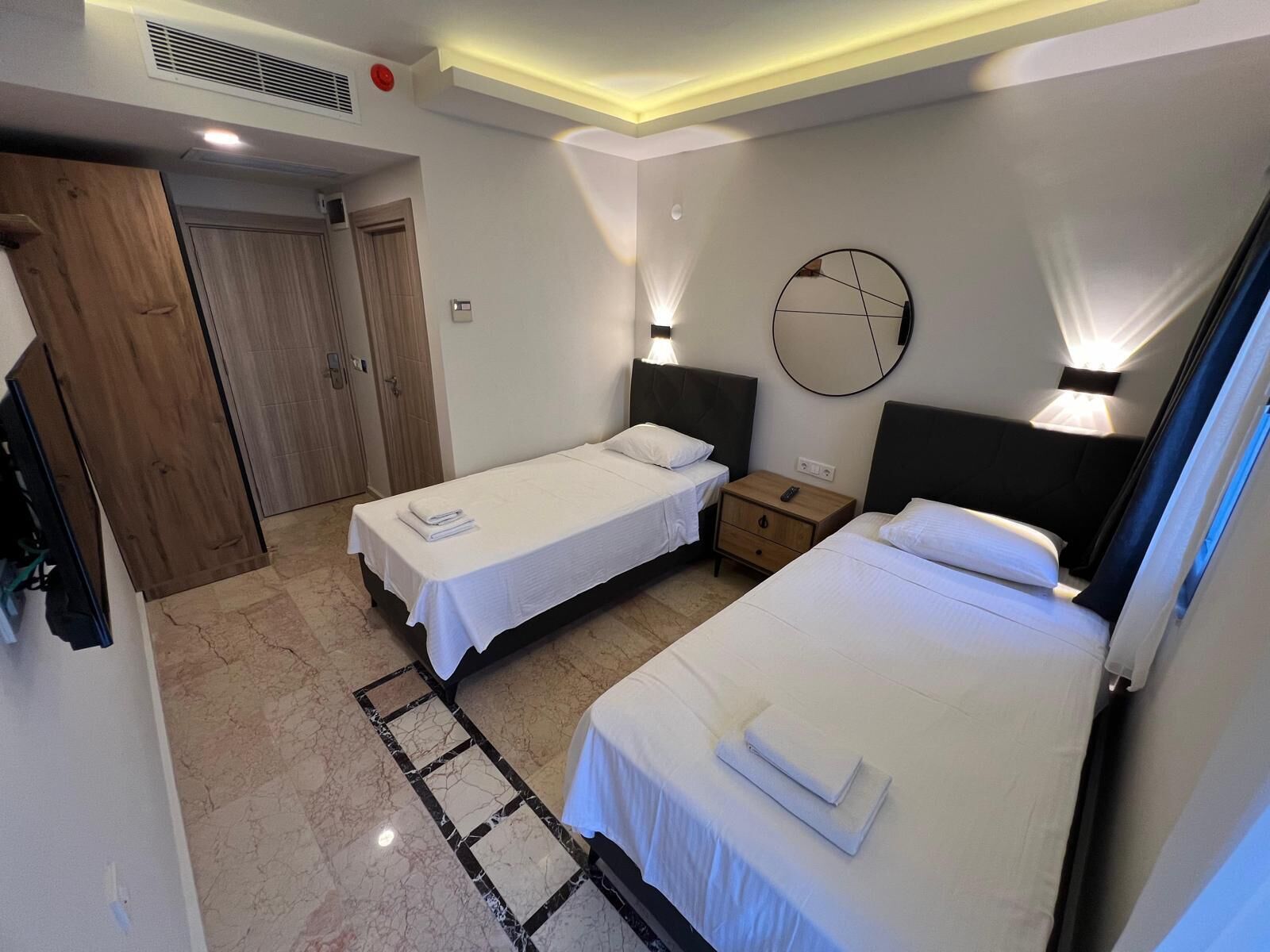 Una habitación de hotel con dos camas individuales, un armario de madera, un espejo redondo y una mesita de noche pequeña con una lámpara.