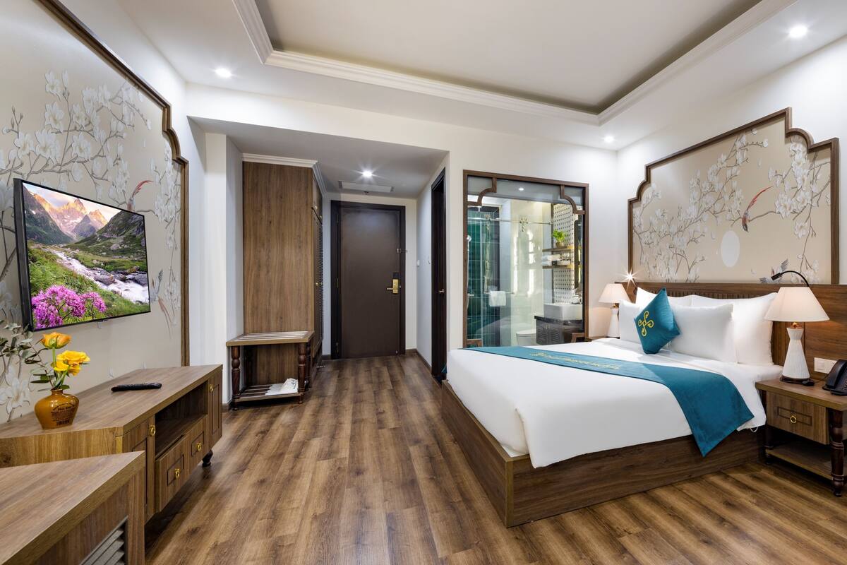 Ein Hotelzimmer mit Holzboden, einem Bett mit weißer Bettwäsche und einem blauen Überwurf, einem Nachttisch mit Lampe, einem an der Wand befestigten Fernseher und einer Holzkommode mit Blumenvase.