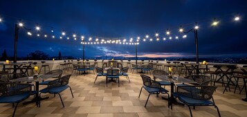 Un espacio de comedor al aire libre con mesas y sillas, luces colgantes y vista a la ciudad al atardecer.
