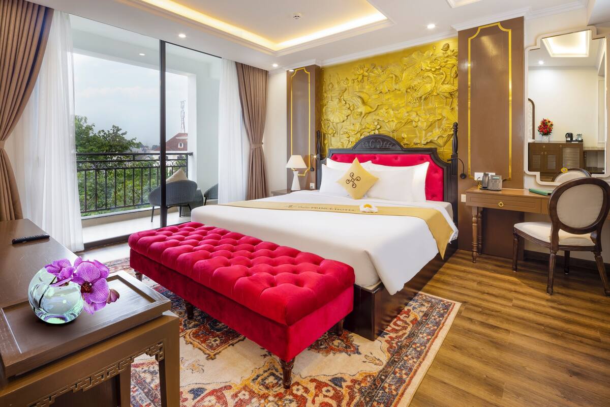 Ein Hotelzimmer mit einem großen Bett, einer roten Bank, einem Schreibtisch und einem Stuhl. Ein Fenster mit Vorhängen bietet einen Blick ins Freie.