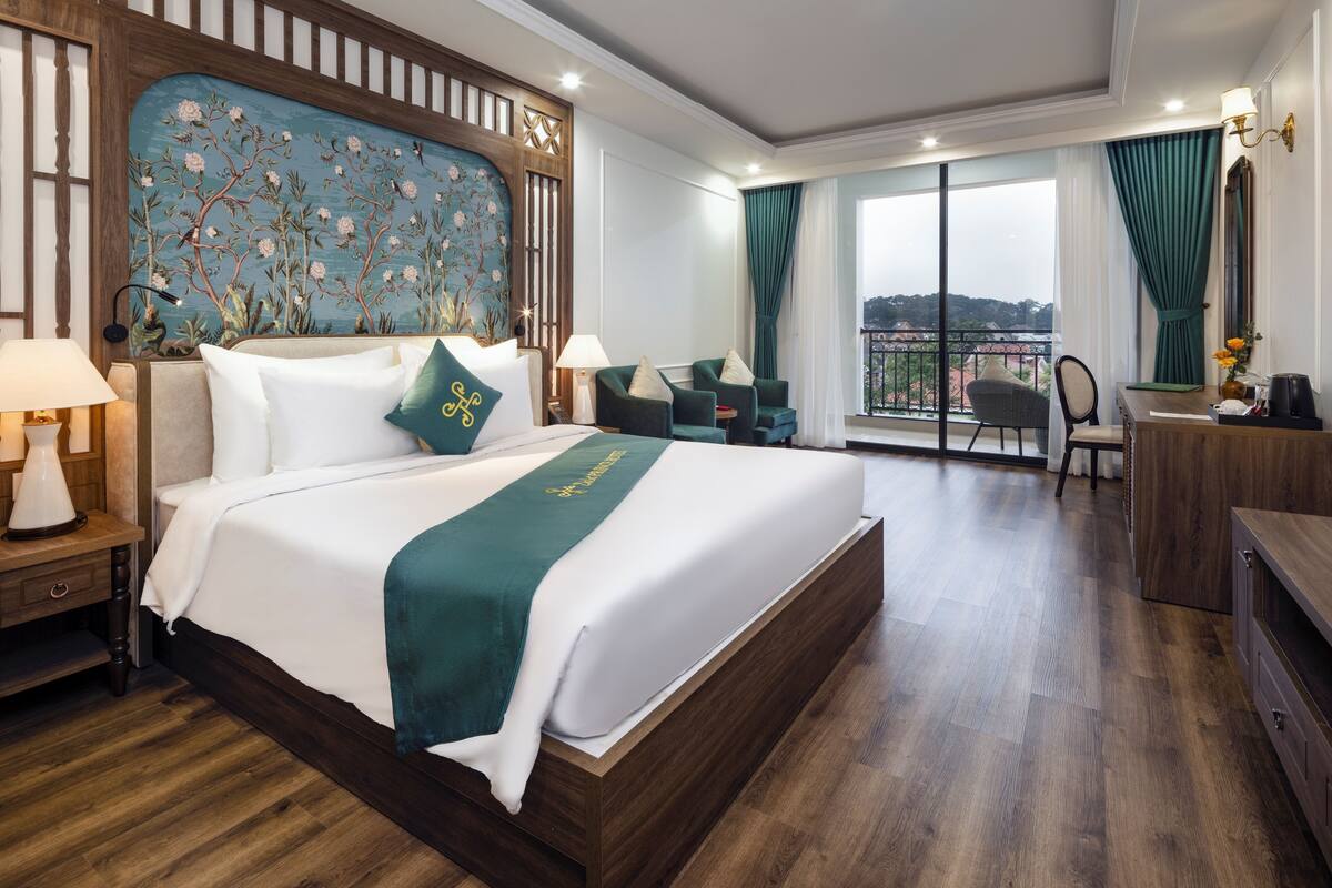 Ein Hotelzimmer mit einem großen Bett, einem Schreibtisch, einem Sessel und einem Balkon mit Blick, sowie einer Wand mit Dekoration.