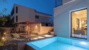 A modern outdoor pool area with lounge chairs and a dining set, adjacent to a building with a glass door leading to a kitchen. - Nisi Living Concept - Kasos Villa (Ialysos)