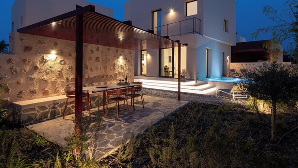 A modern two-story house with a stone exterior, a covered patio with a table and chairs, and a pool area. - Nisi Living Concept - Kasos Villa (Ialysos)