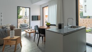 A modern kitchen with a built-in island, a dining area with a table and chairs, a sofa, and a TV mounted on the wall. - Nisi Living Concept - Kasos Villa (Ialysos)