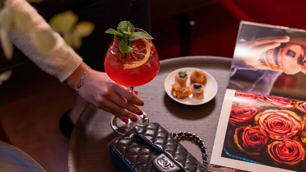 A person holding a cocktail glass with a garnish, a plate of appetizers, a handbag, and a floral print. - Villa-des-Prés (Paris)
