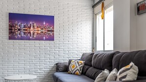 A modern living room with a white brick wall, a cityscape painting, a gray sofa with patterned cushions, and a small round table. - Spacious 'Amplio Canet' Apartment with Shared Pool, Wi-Fi, and Air Conditioning (Canet de Berenguer)