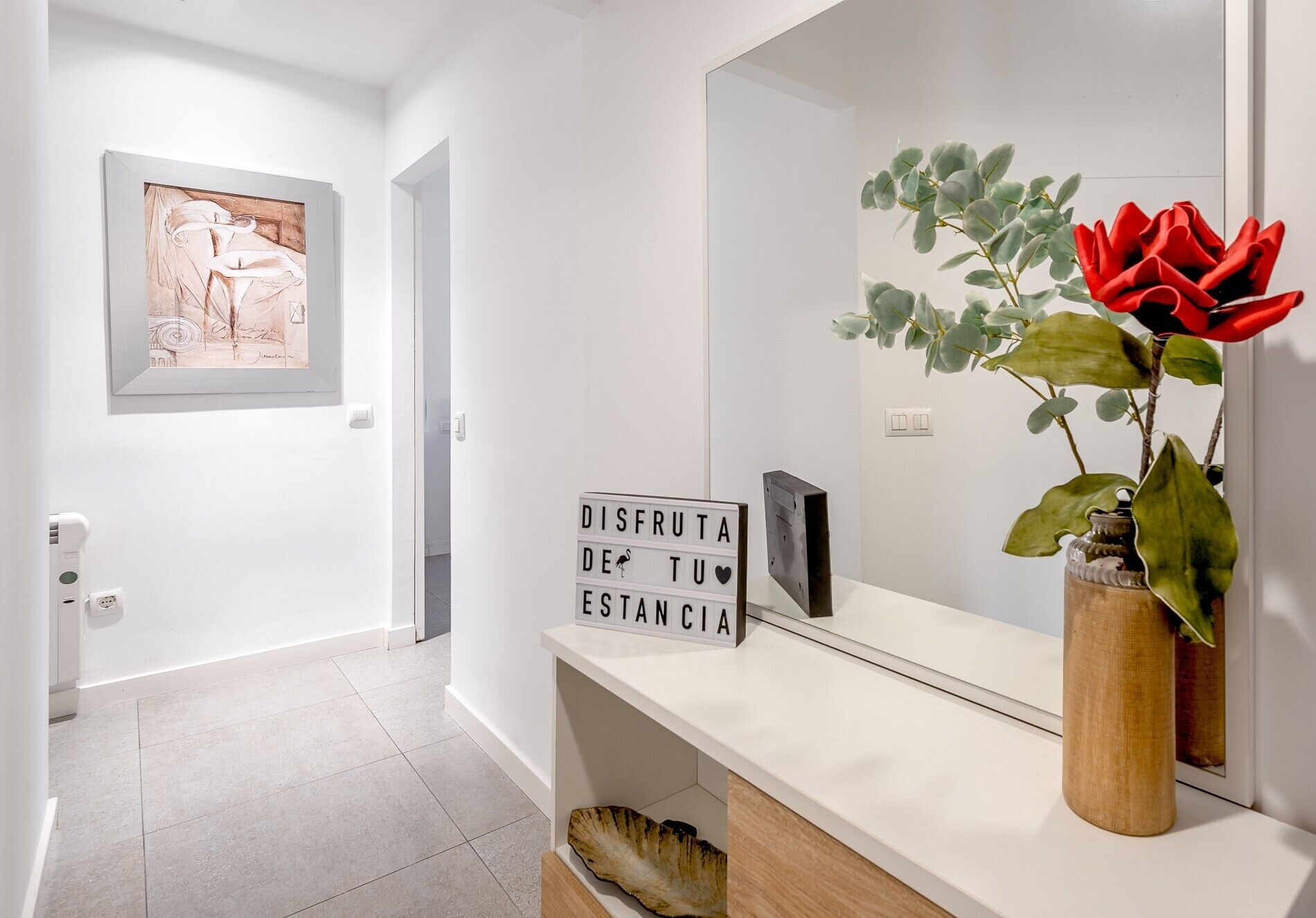 Un couloir avec un mur blanc, une œuvre d’art encadrée, un panneau lumineux, un miroir et une plante en pot ornée de fleurs rouges.