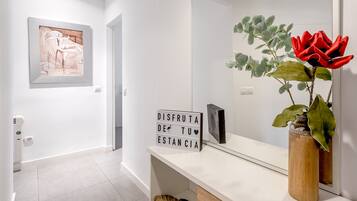 Un couloir avec un mur blanc, une œuvre d’art encadrée, un panneau lumineux, un miroir et une plante en pot ornée de fleurs rouges.