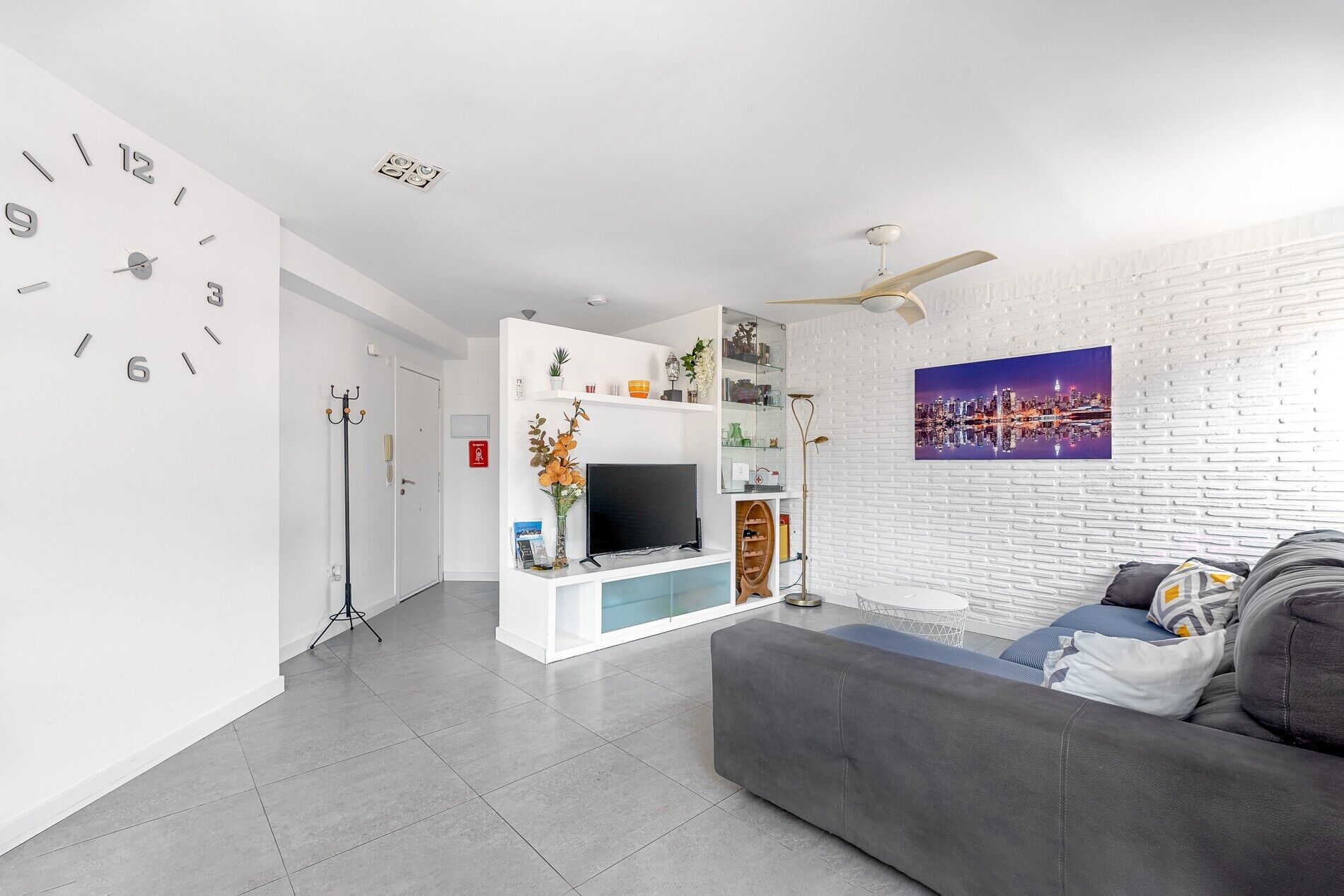 Una sala moderna con televisor de pantalla plana, un sofá gris, ventilador de techo y una pintura de paisaje urbano en la pared.