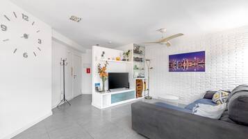 Una sala moderna con televisor de pantalla plana, un sofá gris, ventilador de techo y una pintura de paisaje urbano en la pared.