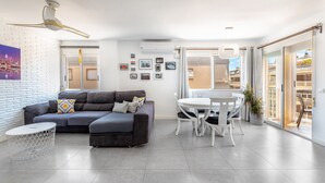 A modern living room with a grey sofa, white dining table, and chairs, a ceiling fan, and a cityscape view through the window. - Spacious 'Amplio Canet' Apartment with Shared Pool, Wi-Fi, and Air Conditioning (Canet de Berenguer)