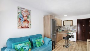 Un salon moderne avec un canapé bleu, des coussins colorés, une cuisine dotée de meubles en bois, un réfrigérateur et un lave-linge.