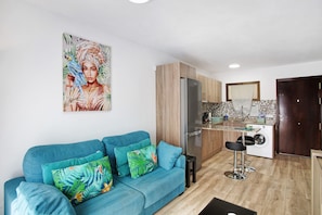 Un salon moderne avec un canapé bleu, des coussins colorés, une cuisine dotée de meubles en bois, un réfrigérateur et un lave-linge.