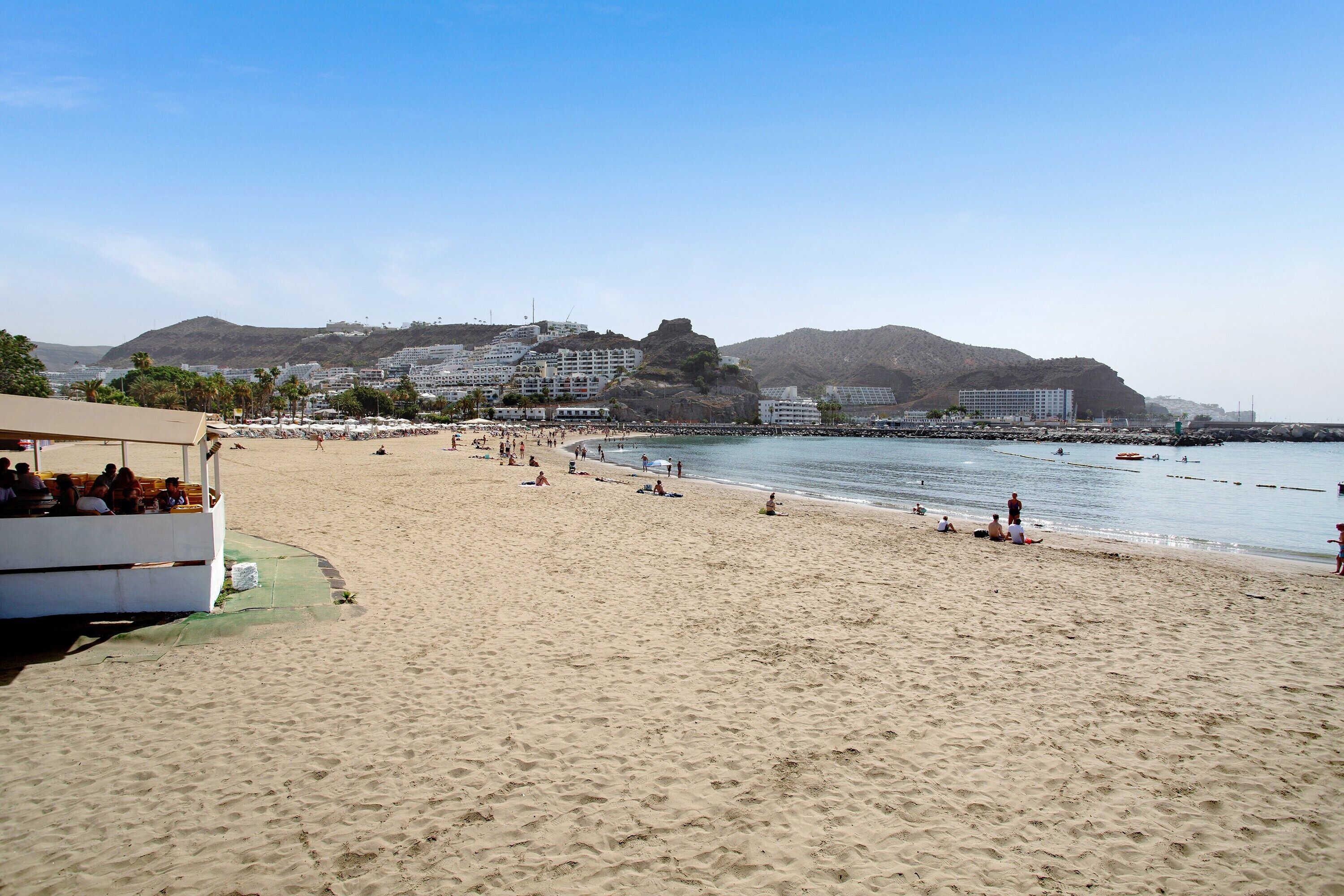 A beach with a white building, a rocky hill, and a clear sky.