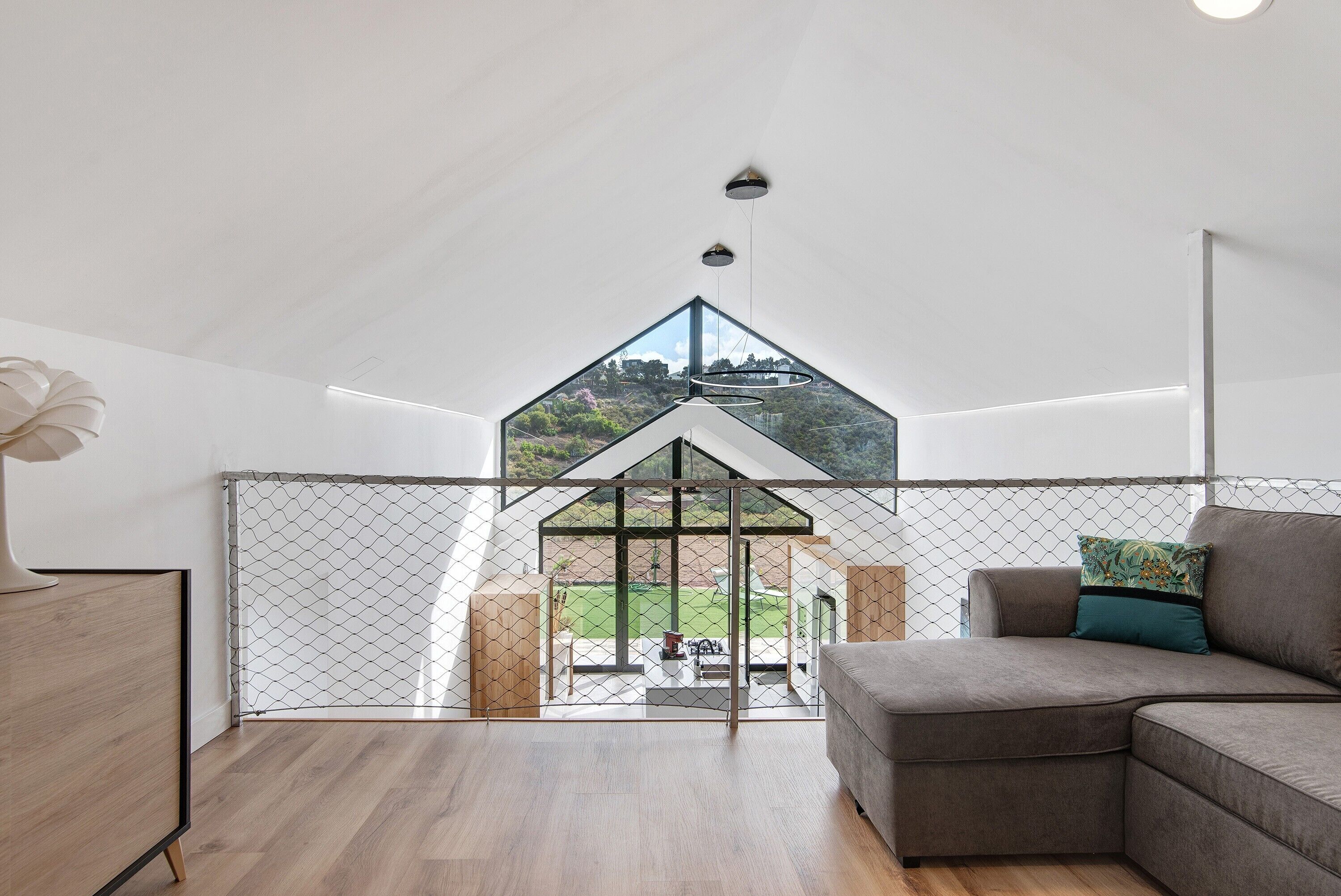 Un intérieur moderne avec un plancher en bois, un canapé d’angle gris, un plafond blanc et une vue dégagée grâce à une cloison en verre.