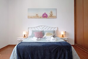 Une chambre à coucher comprenant un lit, deux tables de chevet, une commode, un tableau accroché au mur et une porte en bois.