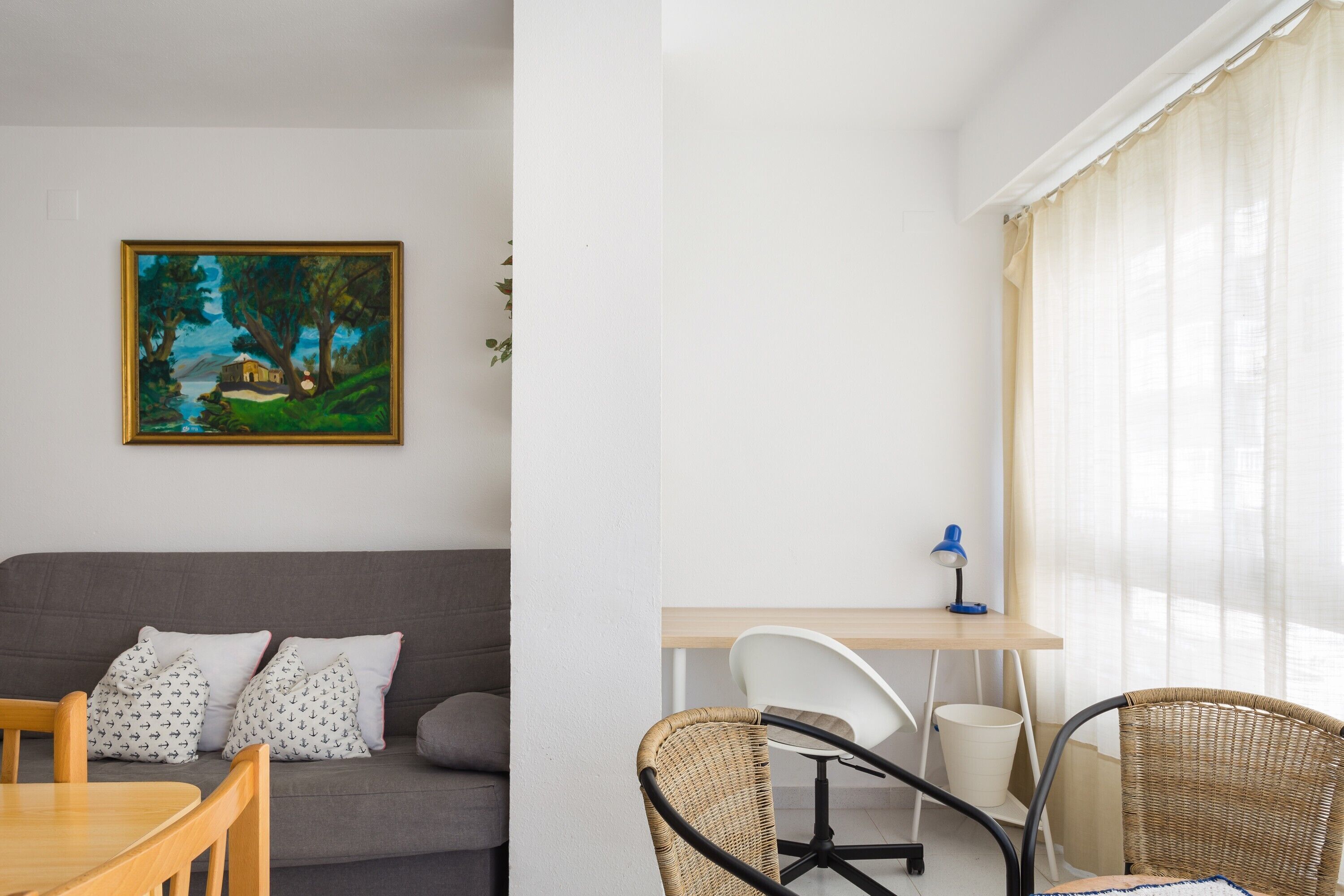 Una sala de estar moderna con un sofá gris, una mesa de comedor, sillas de mimbre, un escritorio con una computadora y un cuadro en la pared.